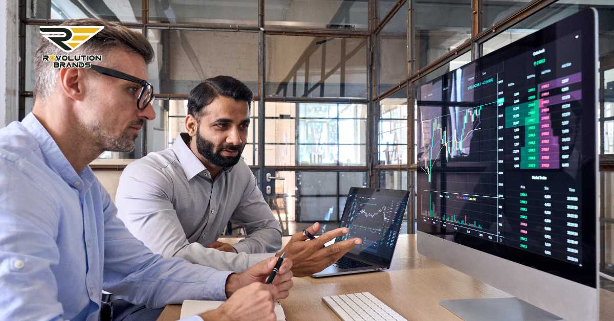 Two professional businessmen analyzing financial data on multiple screens displaying dynamic charts and market trends in a modern office environment. The men, one wearing glasses and a light blue shirt, and the other in a grey shirt, are engaged in a detailed discussion, pointing at the screens and using a laptop for further insights. The R3volution Brands logo in the upper left corner signifies a focus on sophisticated growth strategies for franchise investors.