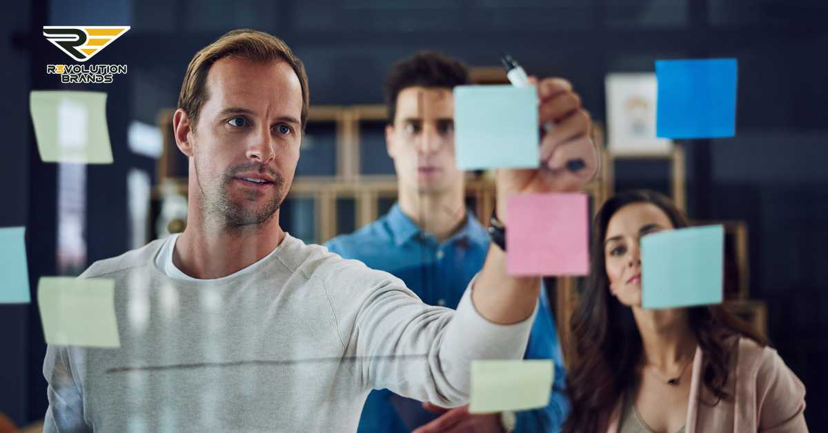 The image captures a collaborative team meeting in a modern office setting, where three team members are engaged in brainstorming using colorful sticky notes on a glass wall. The focus is on a man in the foreground, who appears to be leading the discussion or presenting an idea, as he places a sticky note on the glass. His colleagues, a man and a woman, are also actively participating by adding their own notes to the collection. The R3volution Brands logo in the top left corner ties the activity to the brand's emphasis on strategic planning and team collaboration in the context of building successful franchises. This setting portrays a dynamic and interactive approach to business planning and decision-making.