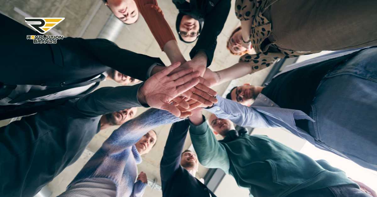 A low-angle view of a diverse group of business professionals putting their hands together in a unified gesture of teamwork and collaboration, viewed from beneath. The circle of people, viewed from this angle, emphasizes unity and the strength of working together. The R3volution Brands logo in the upper left corner aligns the image with themes of community and cooperative success in business."