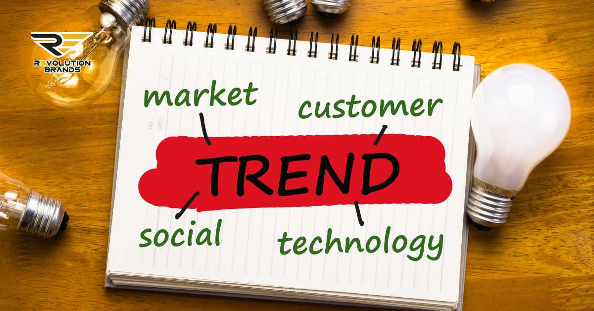 A conceptual image featuring a spiral notebook with the word 'TREND' highlighted in red, surrounded by key business terms like 'market,' 'customer,' 'social,' and 'technology' on a wooden desk. Two light bulbs, one on and one off, symbolize innovative ideas and insights. The R3volution Brands logo in the upper left corner emphasizes the focus on current and emerging business trends centered around customer-centric approaches.