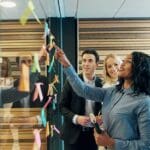 This image shows a group of business professionals engaging in a collaborative activity, likely a strategic planning or brainstorming session, in a modern office setting. They appear to be placing or referring to sticky notes on a glass wall, commonly used for organizing thoughts or assigning tasks during such meetings. The diverse team, including a range of ages and ethnicities, seems engaged and positive, indicating a healthy team dynamic and effective communication. The inclusion of the R3volution Brands logo suggests this scene could be from a company event or workshop focused on innovative business strategies.