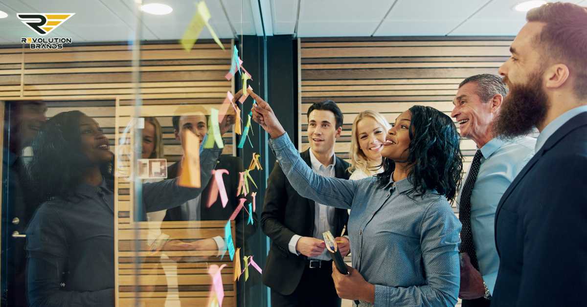 This image shows a group of business professionals engaging in a collaborative activity, likely a strategic planning or brainstorming session, in a modern office setting. They appear to be placing or referring to sticky notes on a glass wall, commonly used for organizing thoughts or assigning tasks during such meetings. The diverse team, including a range of ages and ethnicities, seems engaged and positive, indicating a healthy team dynamic and effective communication. The inclusion of the R3volution Brands logo suggests this scene could be from a company event or workshop focused on innovative business strategies.