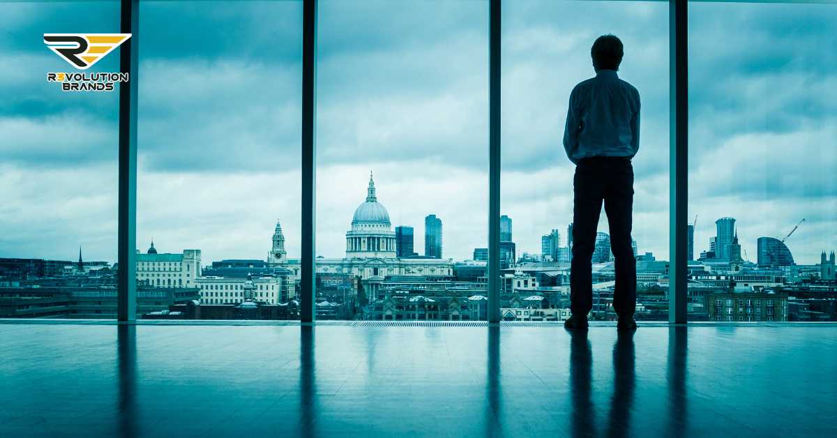 A contemplative man stands in a modern glass-walled office overlooking the London skyline, featuring iconic landmarks like St. Paul's Cathedral. This image captures a moment of reflection, possibly on the broader impact of business practices in the age of social responsibility. The R3volution Brands logo in the upper left corner underscores the company’s commitment to blending business with societal values