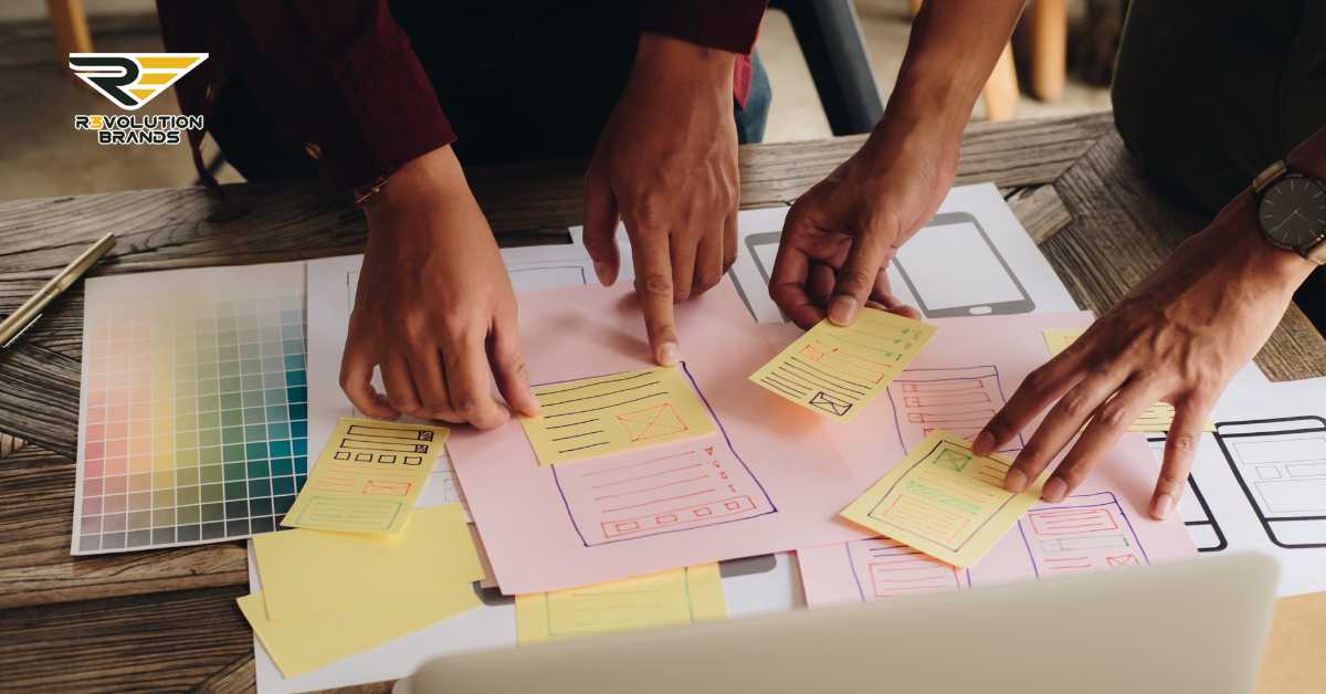 A dynamic team workshop focusing on website and app design, captured at a wood table scattered with color swatches and wireframe layouts. Team members, shown from above, use vibrant sticky notes to collaboratively plan and refine user interface elements. The R3volution Brands logo in the upper left corner aligns with the theme of innovation and adaptive training strategies in franchising.