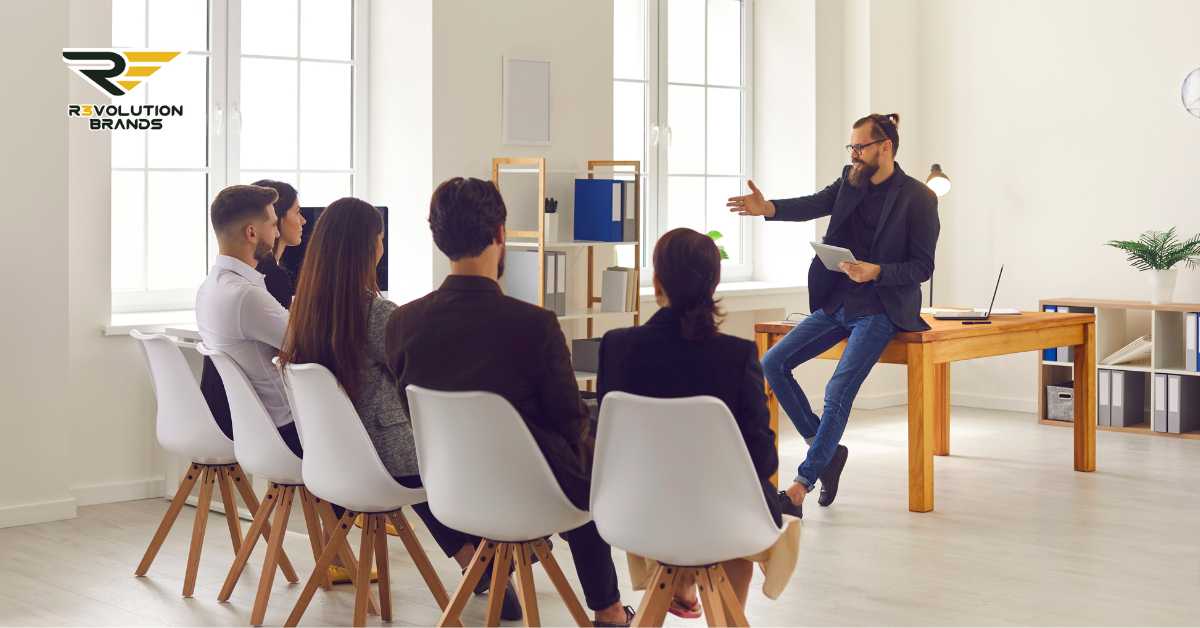 A professional training session at R3volution Brands, featuring a male trainer in casual business attire passionately discussing key points with a diverse group of attentive participants seated in white modern chairs. The bright, spacious office environment with large windows enhances the learning atmosphere. The R3volution Brands logo in the upper left corner emphasizes the company’s commitment to training and developing franchise growth strategies.