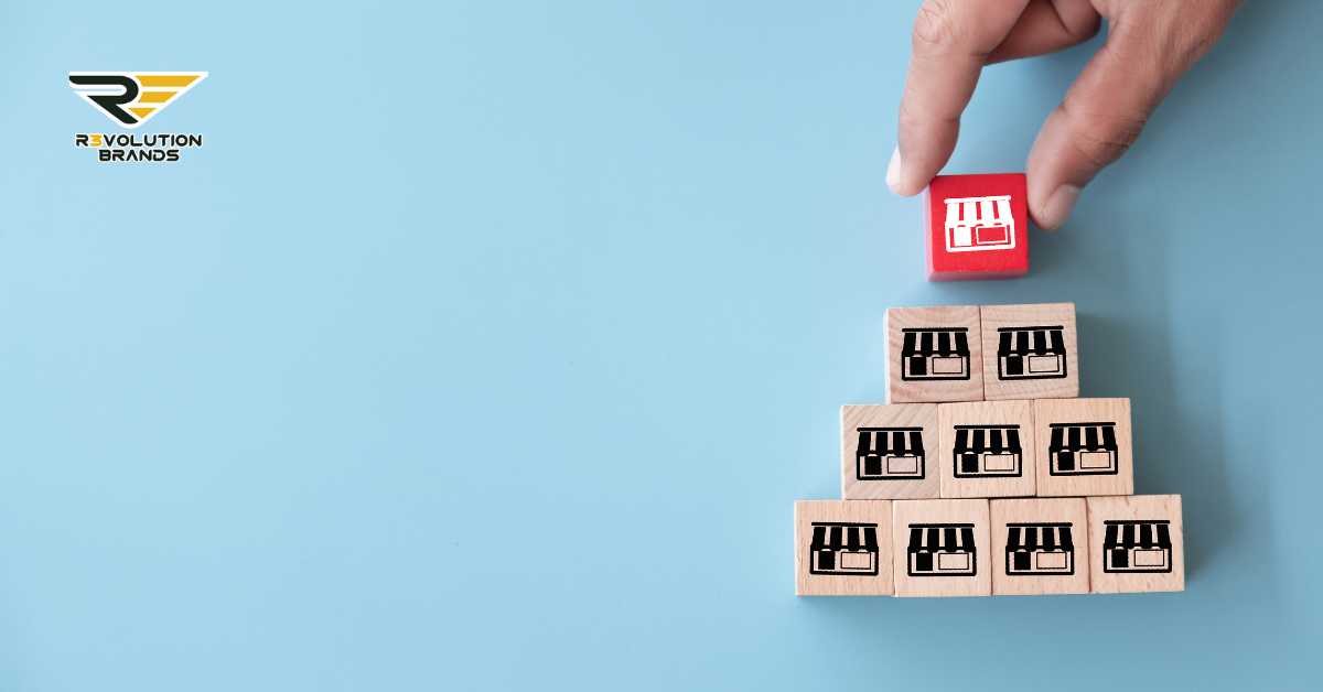 A conceptual image of a hand placing the final red block, with a store icon, atop a pyramid made of wooden blocks each featuring a black store icon, against a light blue background. This visual metaphor represents the strategic building of a franchise system, with the red block symbolizing the culmination or achievement of a strategic goal. The R3volution Brands logo in the upper left corner underscores the focus on innovative growth tactics in franchising.