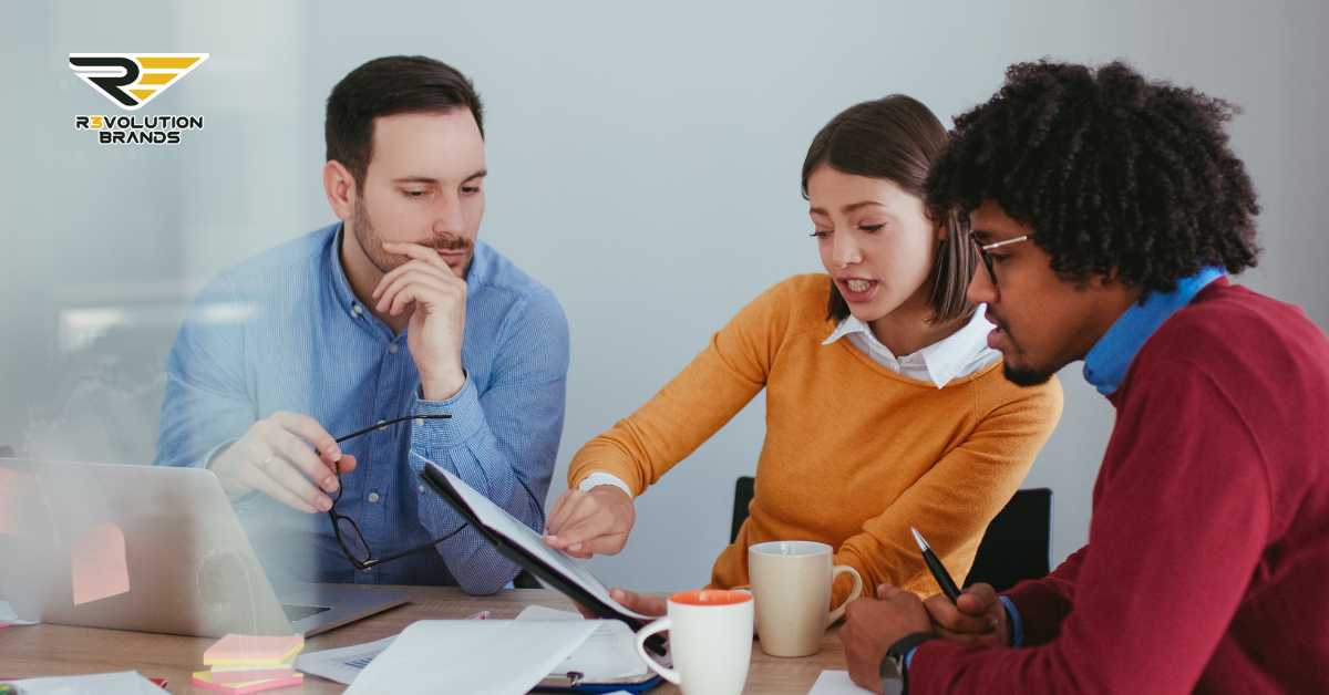 Three colleagues collaborate at a meeting in a modern office setting, displaying the R3volution Brands logo in the upper left corner. A man in a blue shirt attentively listens while holding a pen, a woman in a yellow sweater actively explains a point with a digital tablet in hand, and another man in a red sweater takes notes. They sit around a table with coffee mugs and laptops, indicating a focused work environment.