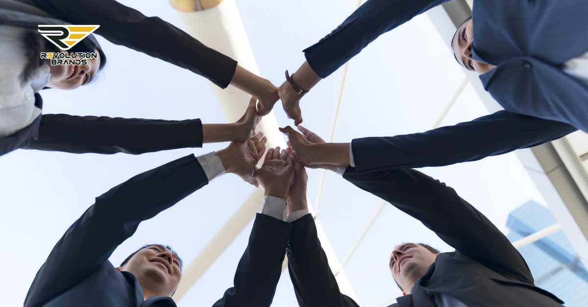 An uplifting view from below of a diverse team of business professionals in a circle, hands joined together in a unified gesture that symbolizes teamwork and collaboration. The team is dressed in professional attire, set against a backdrop of a modern architectural structure and clear sky, reflecting a positive, collaborative work environment. The R3volution Brands logo in the upper left corner accentuates the brand's focus on unity and collective success in the franchise industry.