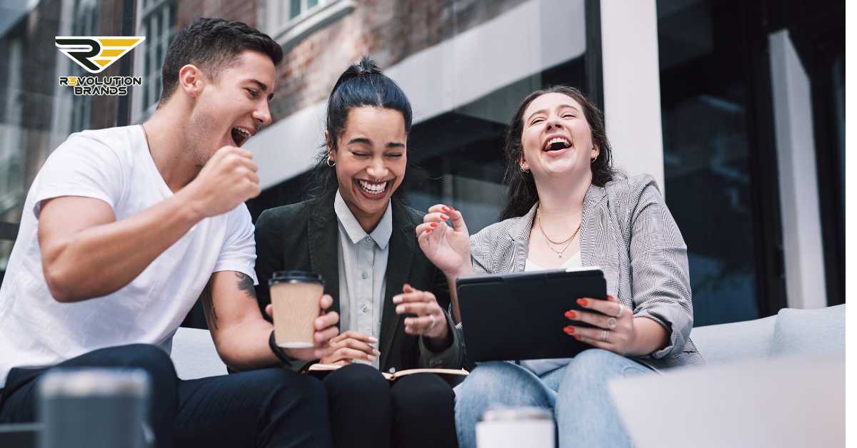 Three young, diverse entrepreneurs share a joyful moment while looking at a tablet outdoors. One man, fist pumped in excitement, celebrates a successful venture while the two women laugh and smile, one holding a coffee. They are casually dressed, suggesting a relaxed yet productive meeting environment. The R3volution Brands logo in the upper left corner highlights the company’s focus on nurturing passionate and profitable franchise investments.